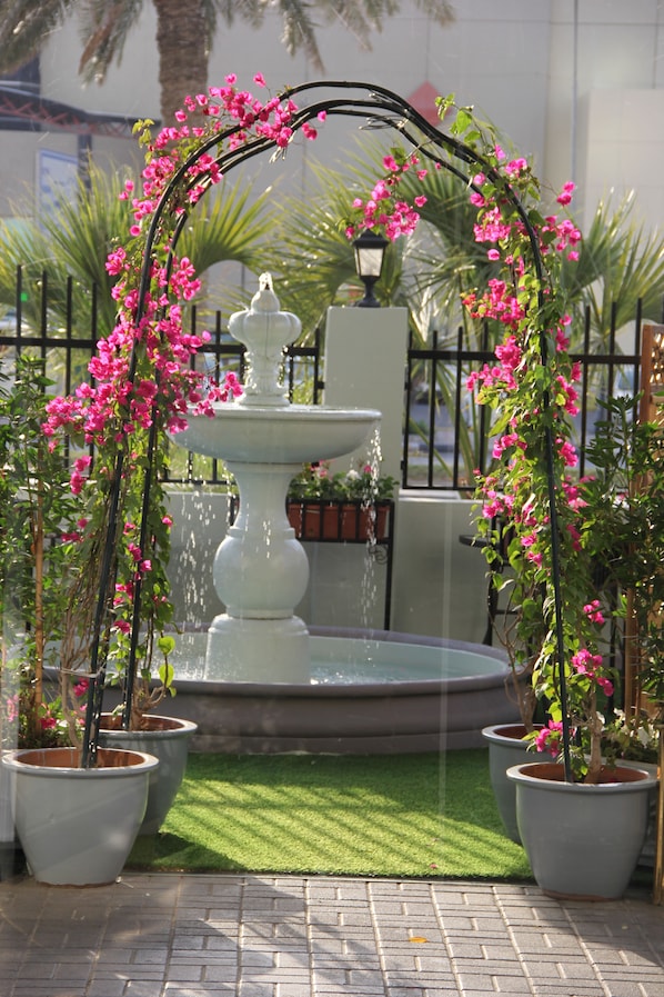 A fountain with a white pedestal, surrounded by pink flowering plants and potted trees, set against a brick pavement and a metal fence.