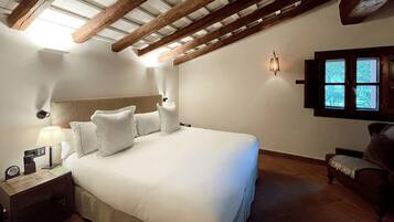 A bedroom with a large bed, wooden beams on the ceiling, a nightstand with a lamp, and a window with a view of trees.