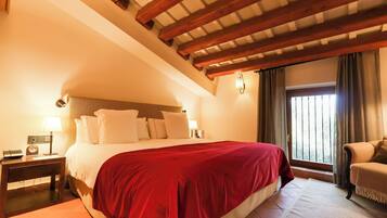 Een slaapkamer met een groot bed, houten balken in het plafond, een nachtkastje met een lamp, een stoel en een raam met gordijnen.