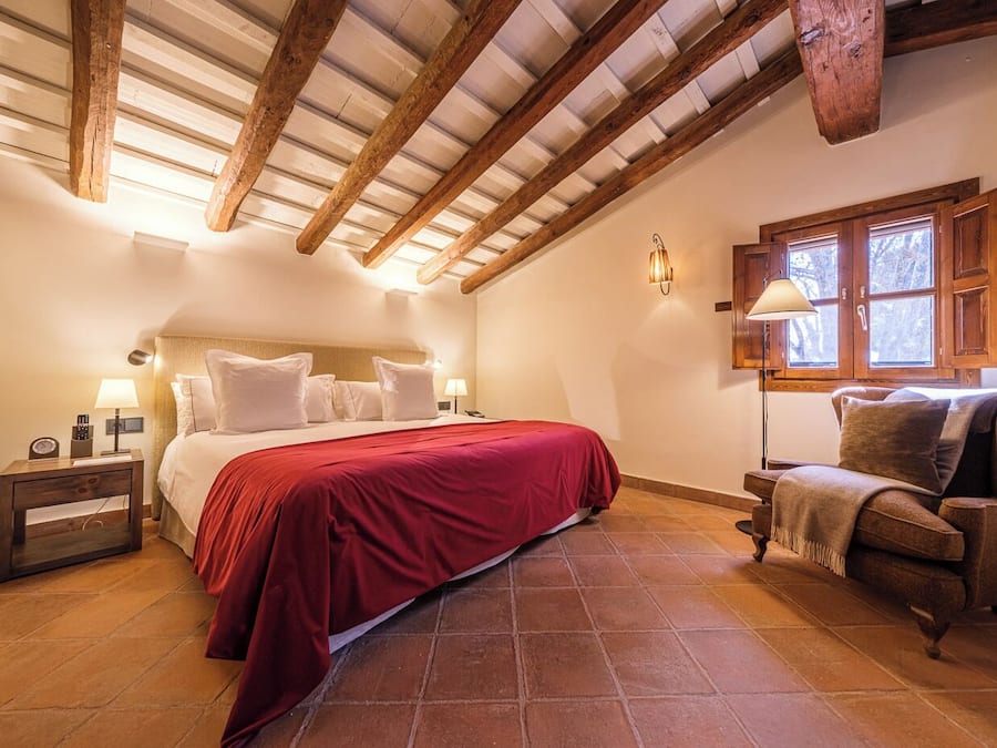 Un dormitorio con una cama grande, vigas de madera en el techo, una mesita de noche, una silla y una ventana con vistas.