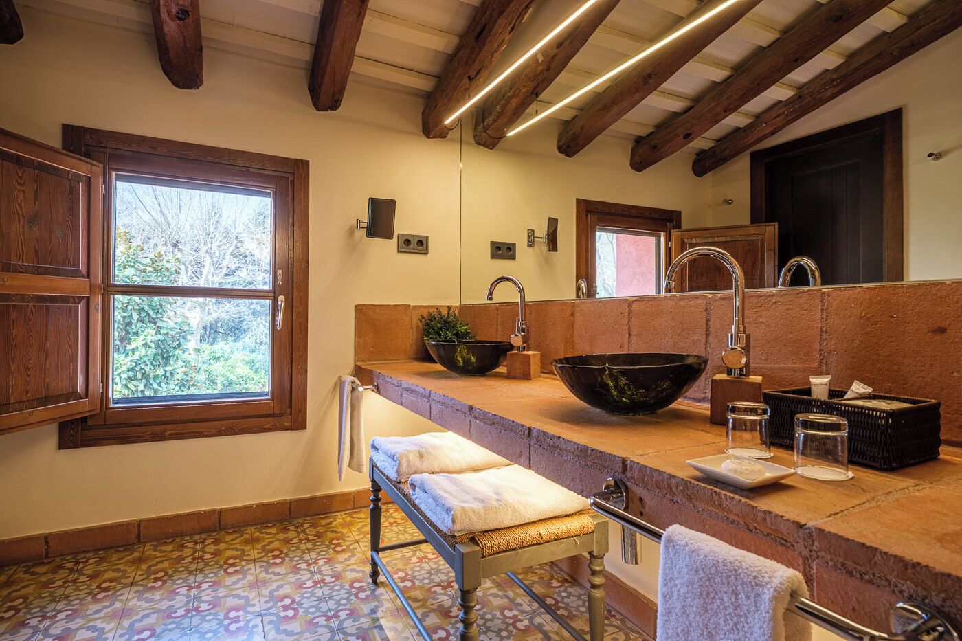 A bathroom with a large window, wooden beams, a sink with a bowl, and a bench.