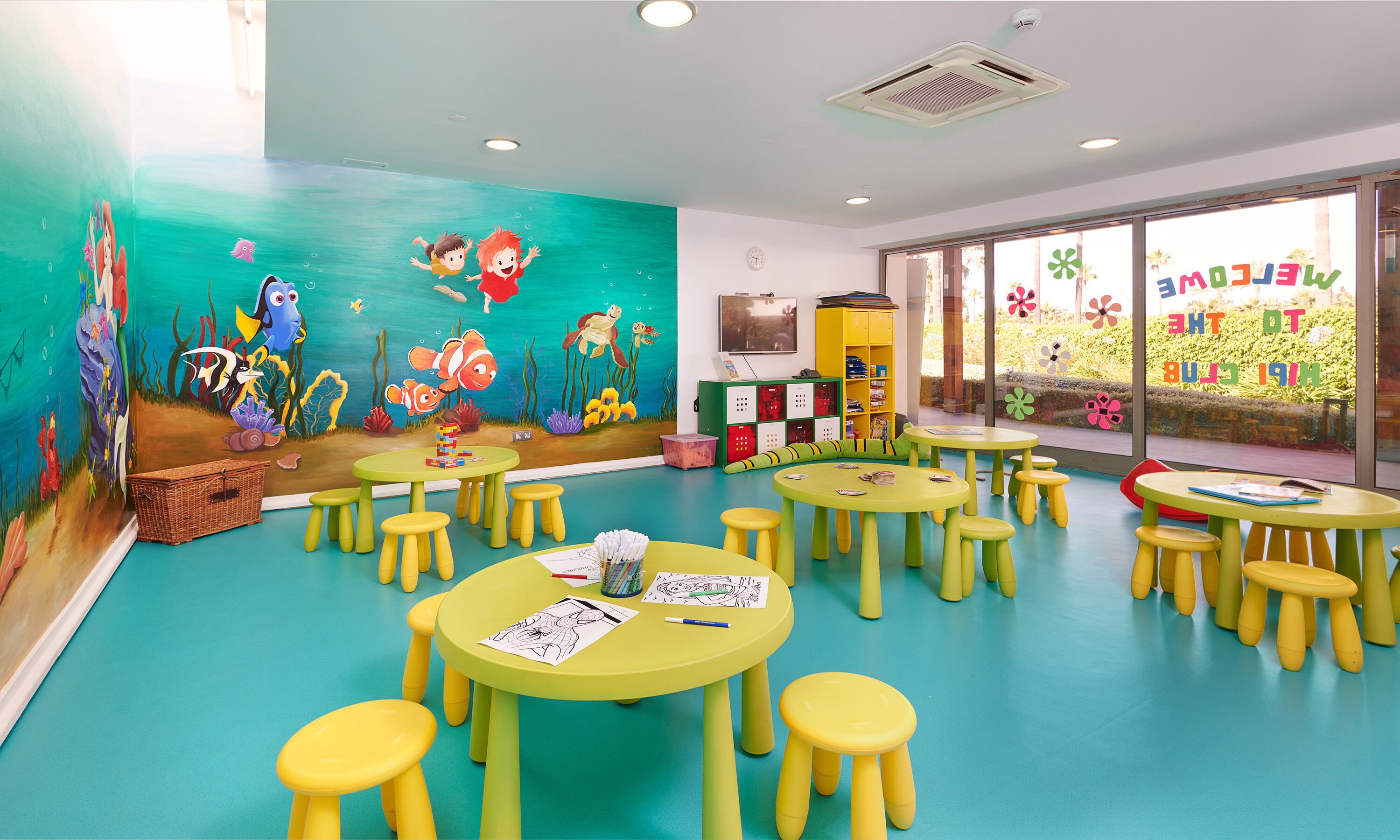 Un cuarto de juegos infantil con murales coloridos, varias mesas y taburetes, y un ventanal con vista a zonas verdes.