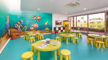 Un cuarto de juegos infantil con murales coloridos, varias mesas y taburetes, y un ventanal con vista a zonas verdes.