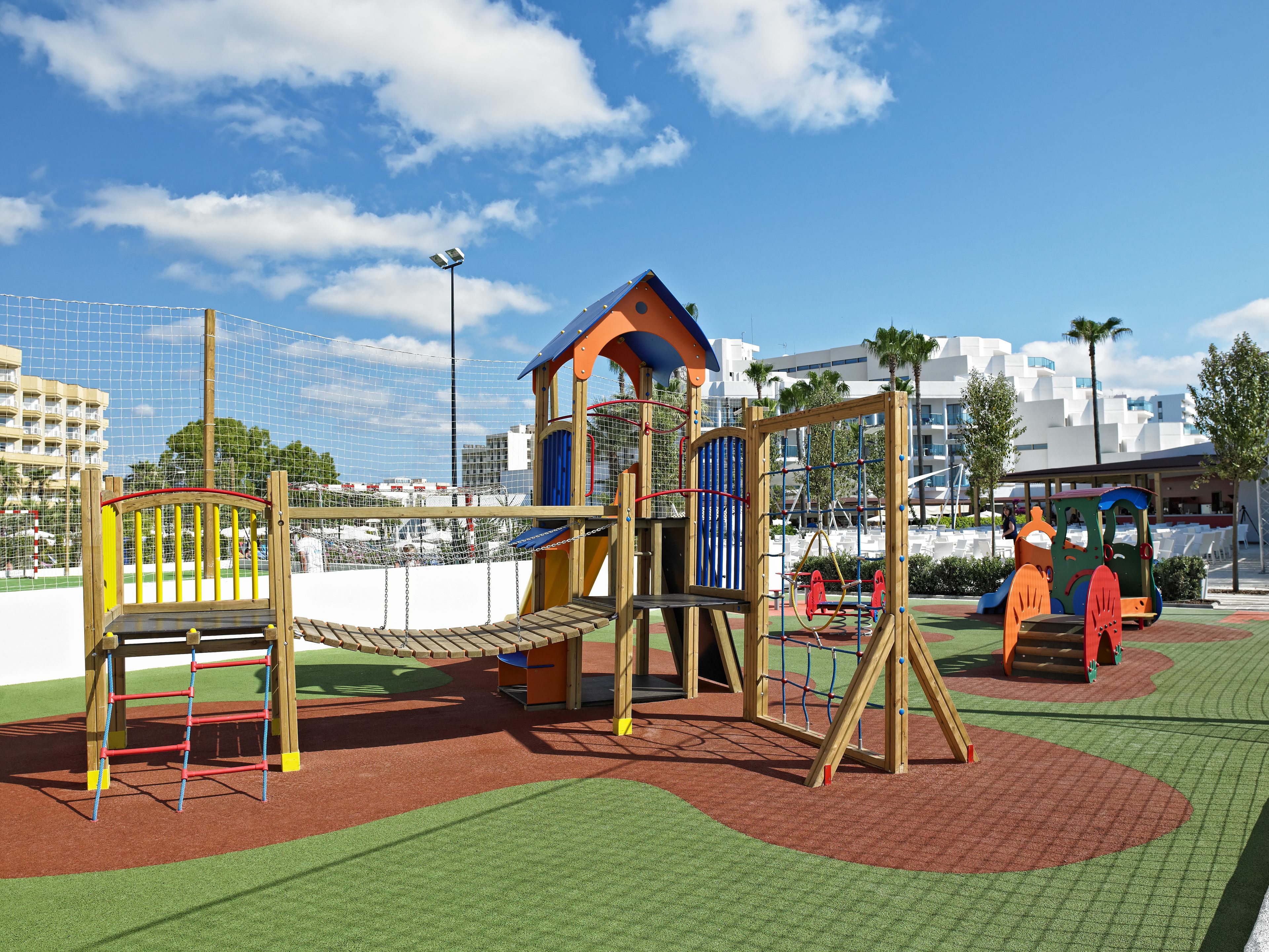 A playground with various play structures, surrounded by a safety net, and a building with white balconies in the background.