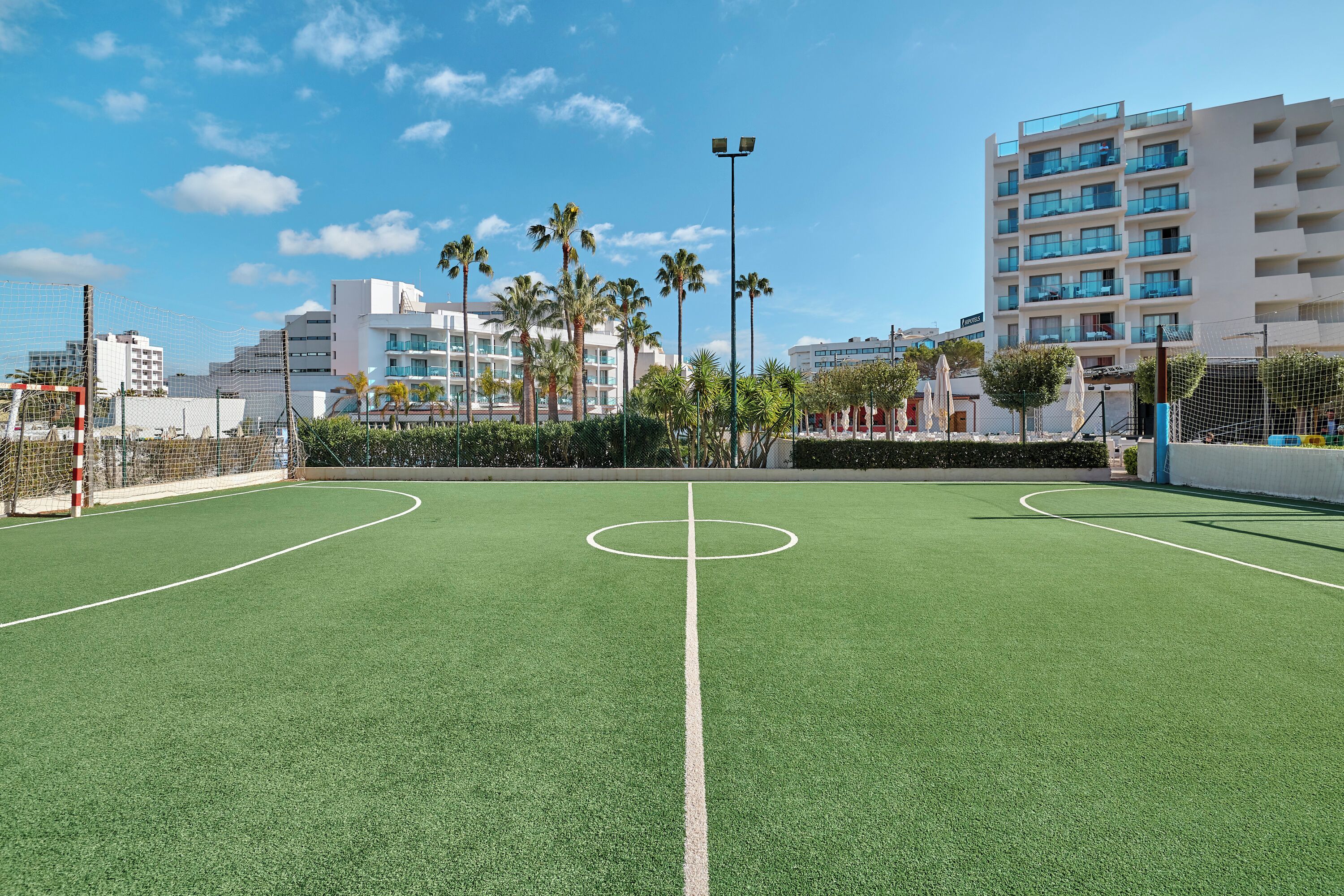 A soccer field with goalposts and a net, surrounded by palm trees and buildings.