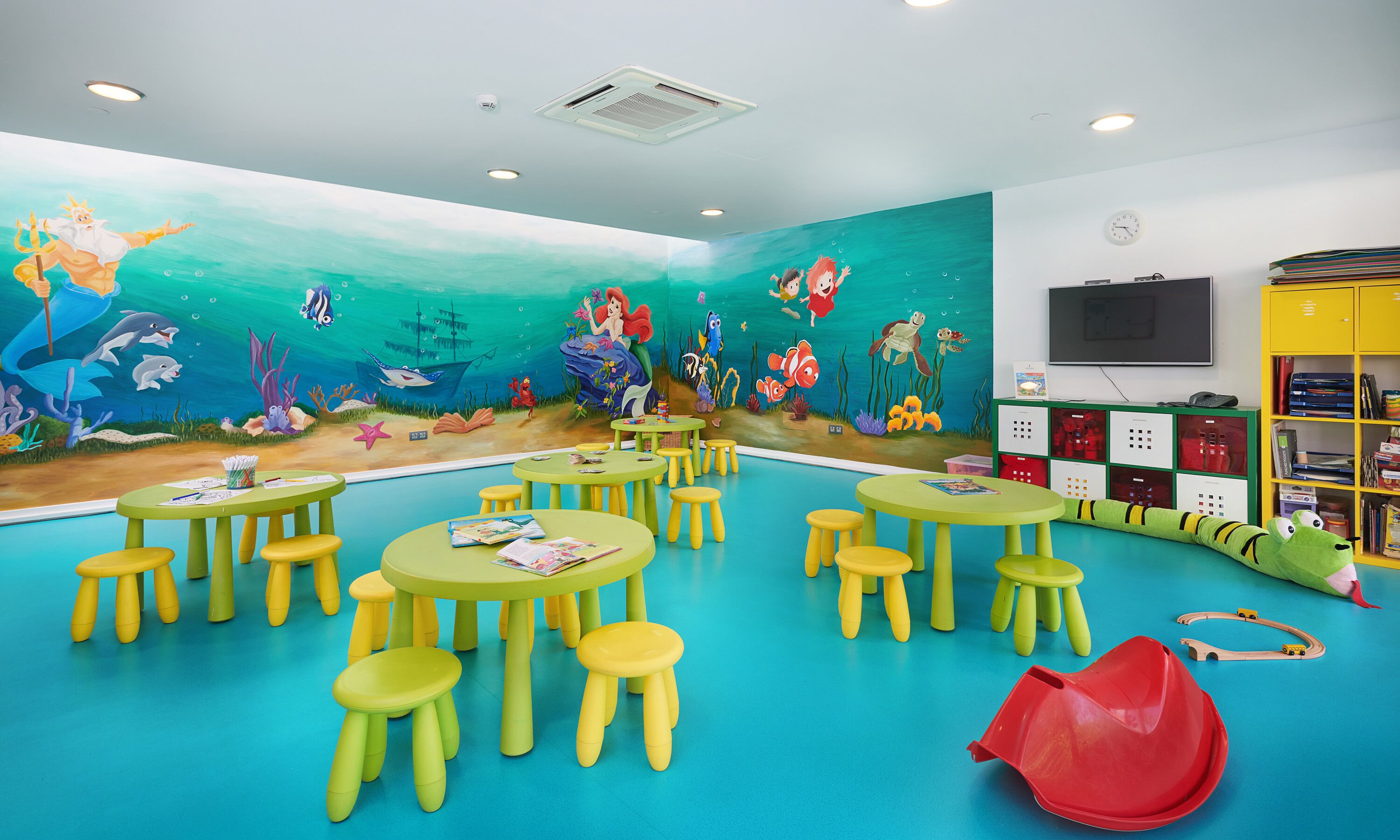 A children's playroom with a mural of an underwater scene, yellow tables and stools, and a bookshelf with toys.