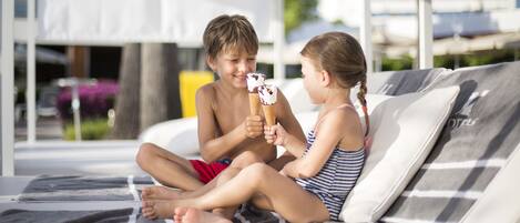 Two children sitting on a lounge chair by a poolside, holding ice cream cones.