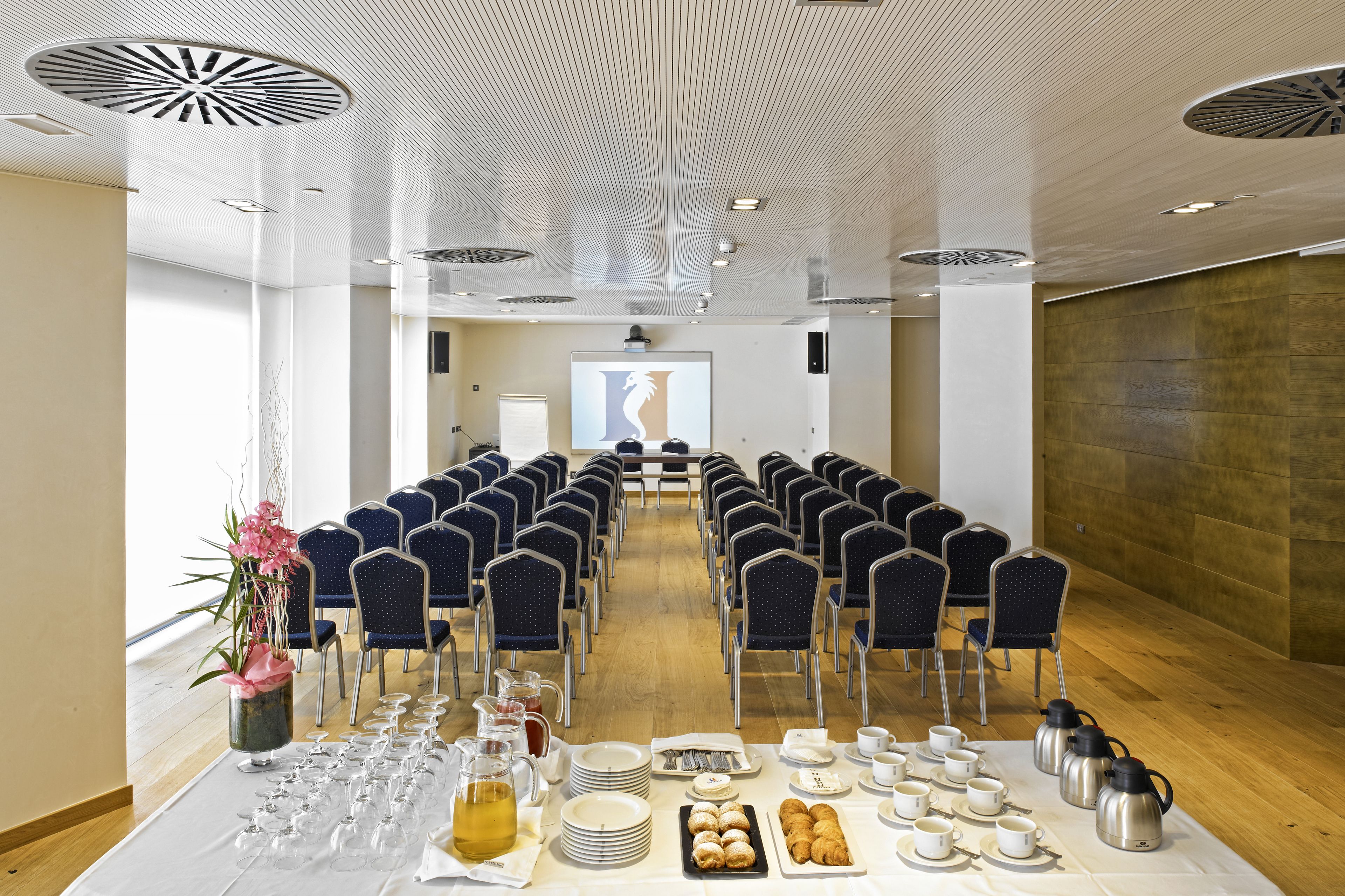 Un salón de conferencias con filas de sillas, una pantalla de proyector y una mesa de buffet con comida y bebidas.