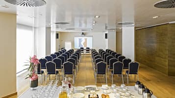 Un salón de conferencias con filas de sillas, una pantalla de proyector y una mesa de buffet con comida y bebidas.