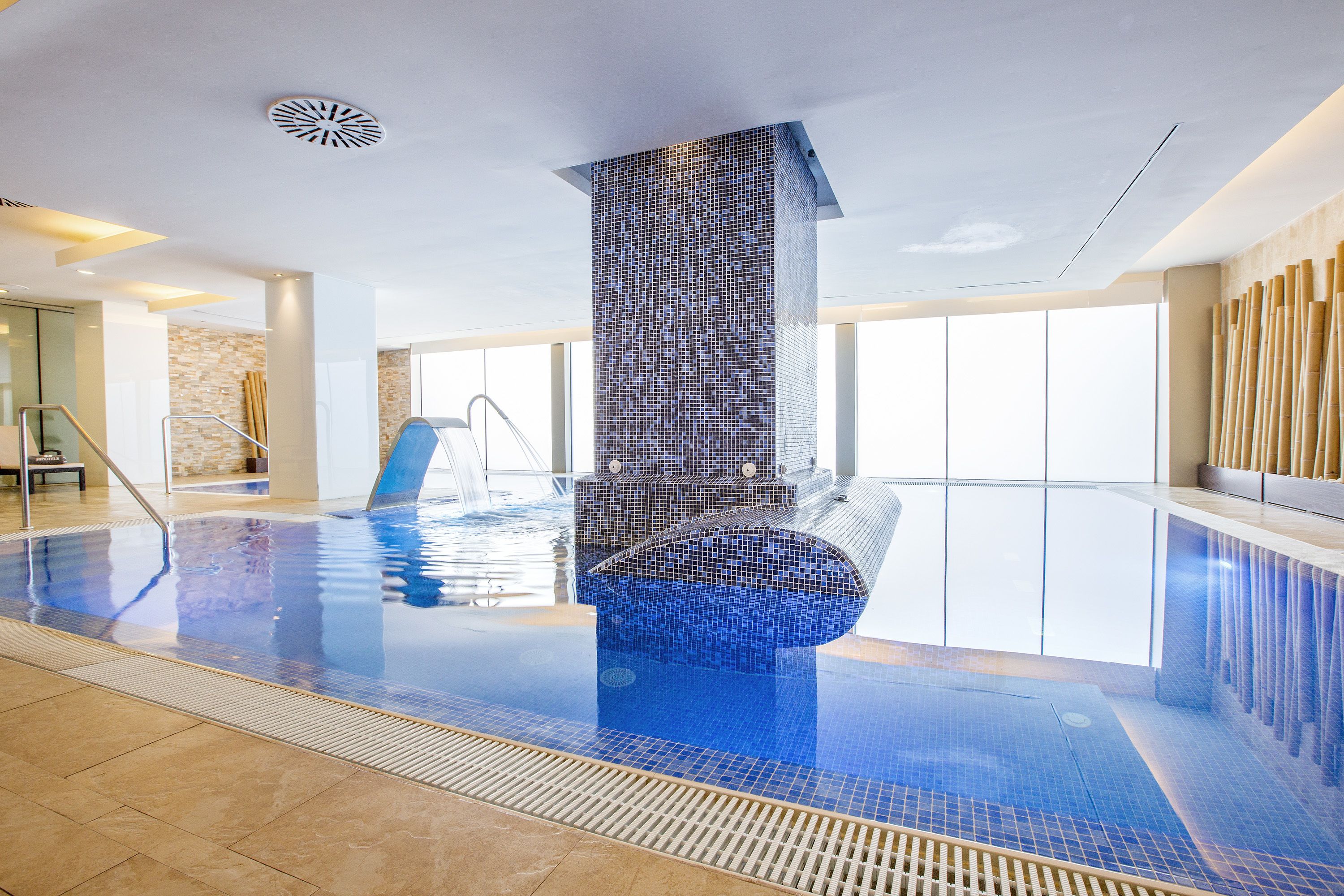 A modern indoor swimming pool with clear water, a tiled wall, and a glass balustrade.