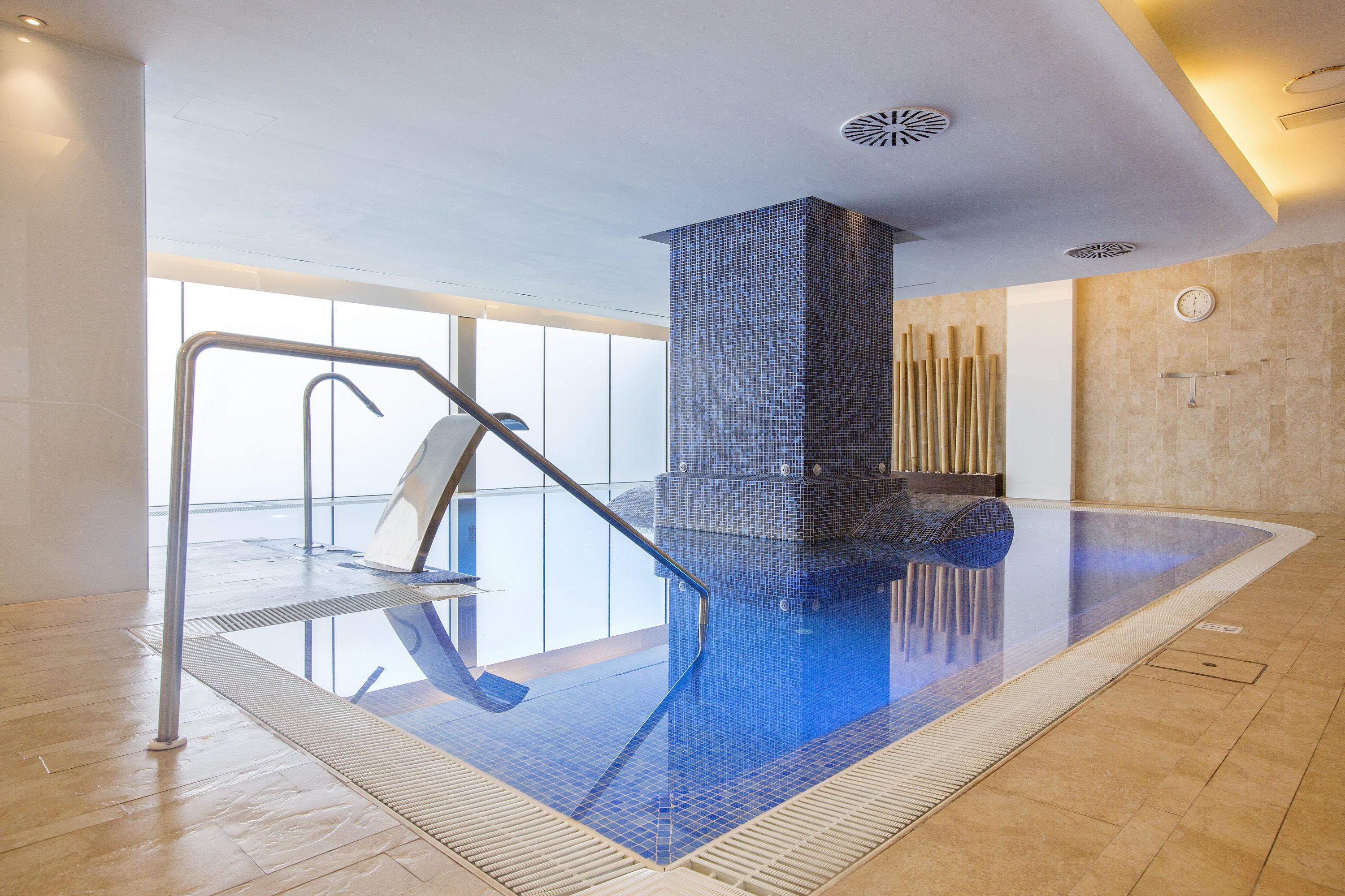 A modern indoor swimming pool with a clear glass floor, a metal ladder, and a tiled wall.