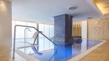 A modern indoor swimming pool with a clear glass floor, a metal ladder, and a tiled wall.