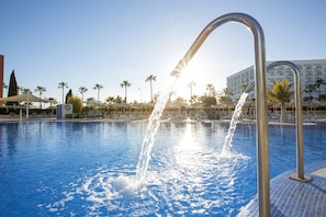 A swimming pool with water jets, surrounded by lounge chairs and palm trees, with a hotel in the background.
