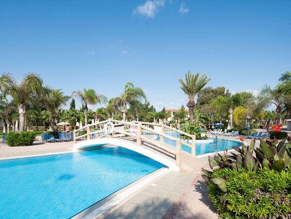 A pool area with a wooden bridge, lounge chairs, and palm trees. - Nelia Sea Breeze (Ayia Napa)