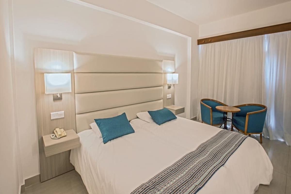 Una habitación de hotel con una cama grande, dos mesitas de noche, un escritorio con sillas y una ventana con cortinas.