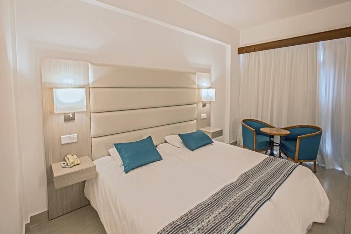 Ein Hotelzimmer mit einem großen Bett, zwei Nachttischen, einem Schreibtisch mit Stühlen und einem Fenster mit Vorhängen.