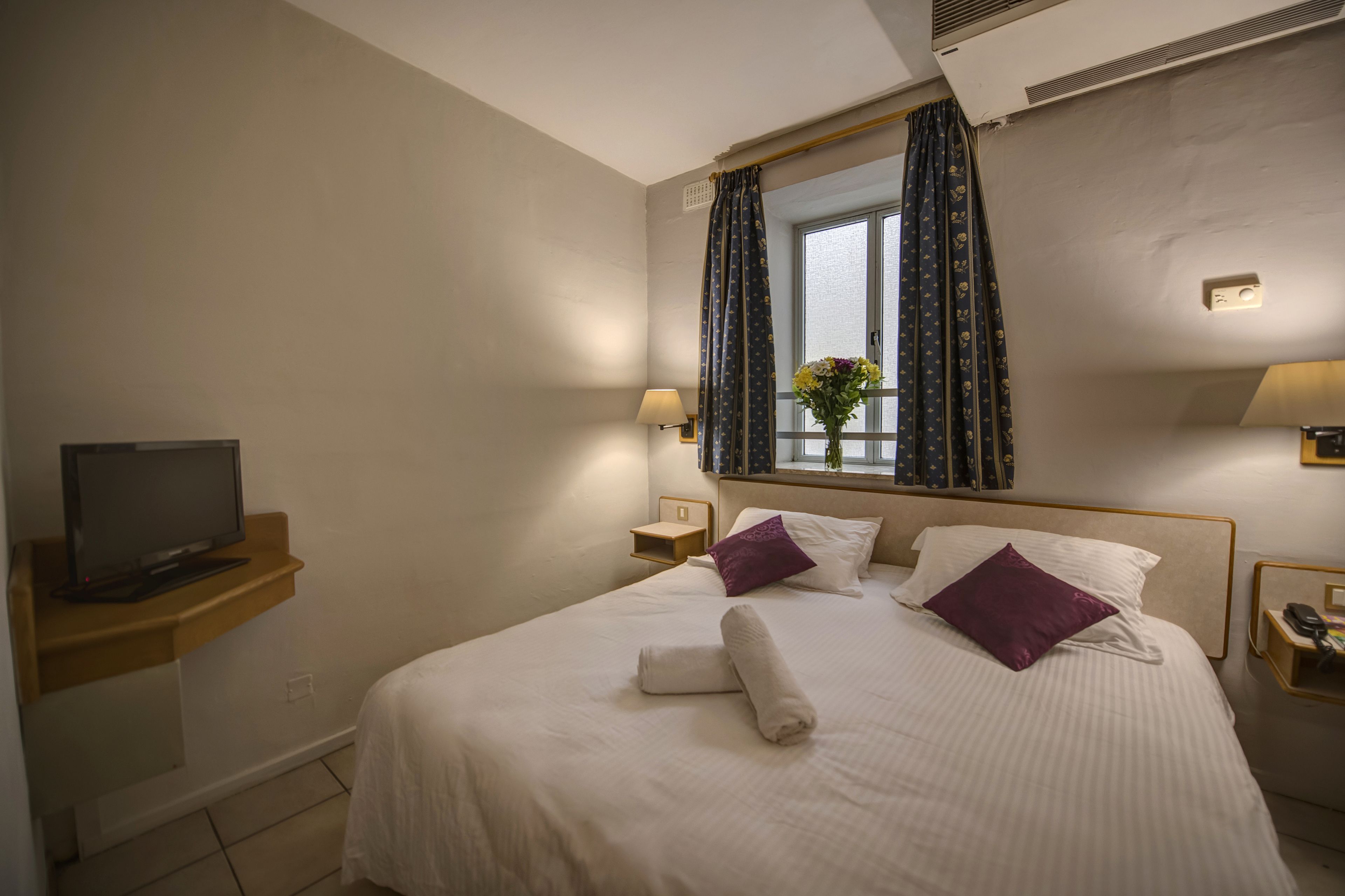A hotel room with a bed, a TV on a stand, a window with curtains, and a nightstand with a lamp.