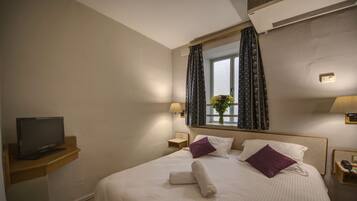 Habitación de hotel con cama, televisor en un soporte, ventana con cortinas y mesita de noche con lámpara.