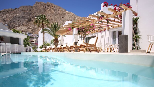 Un espace piscine doté de chaises longues et d’une pergola ornée de fleurs, le tout entouré de bâtiments blancs et avec une montagne en toile de fond.