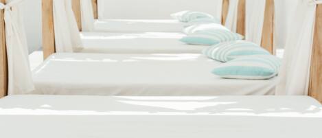 A row of four single beds with white linens and light blue striped pillows, under a canopy of white drapes.