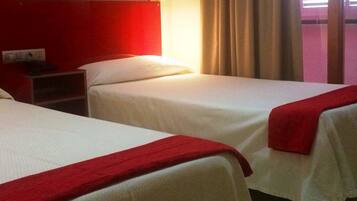 Habitación de hotel con dos camas, cabecera roja, mesita de noche, lámpara de pared y ventana con cortinas.