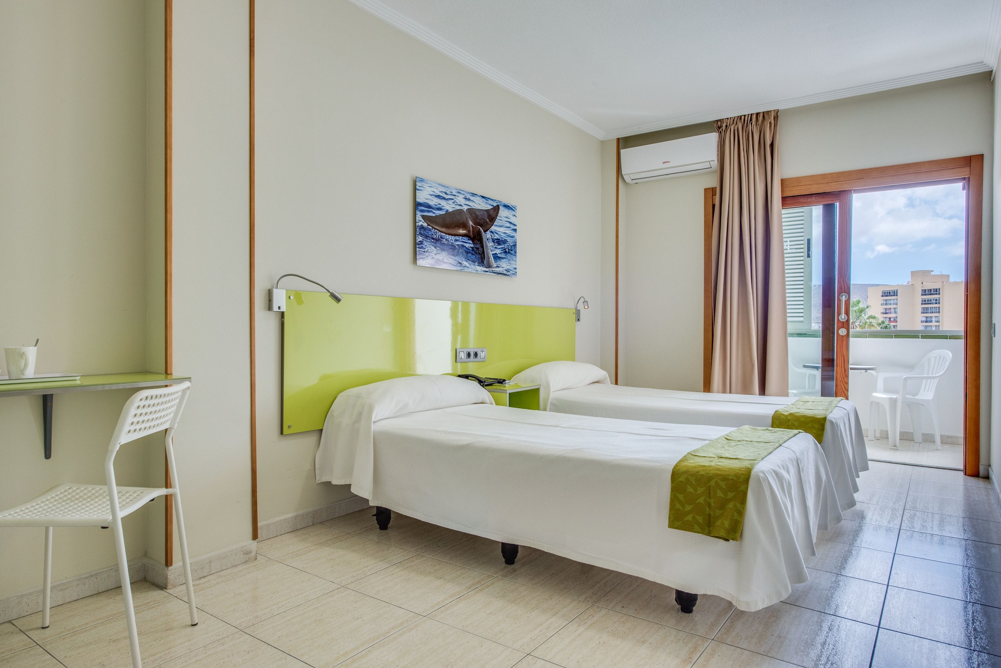 A hotel room with two beds, a desk, a chair, and a window with a view of buildings.