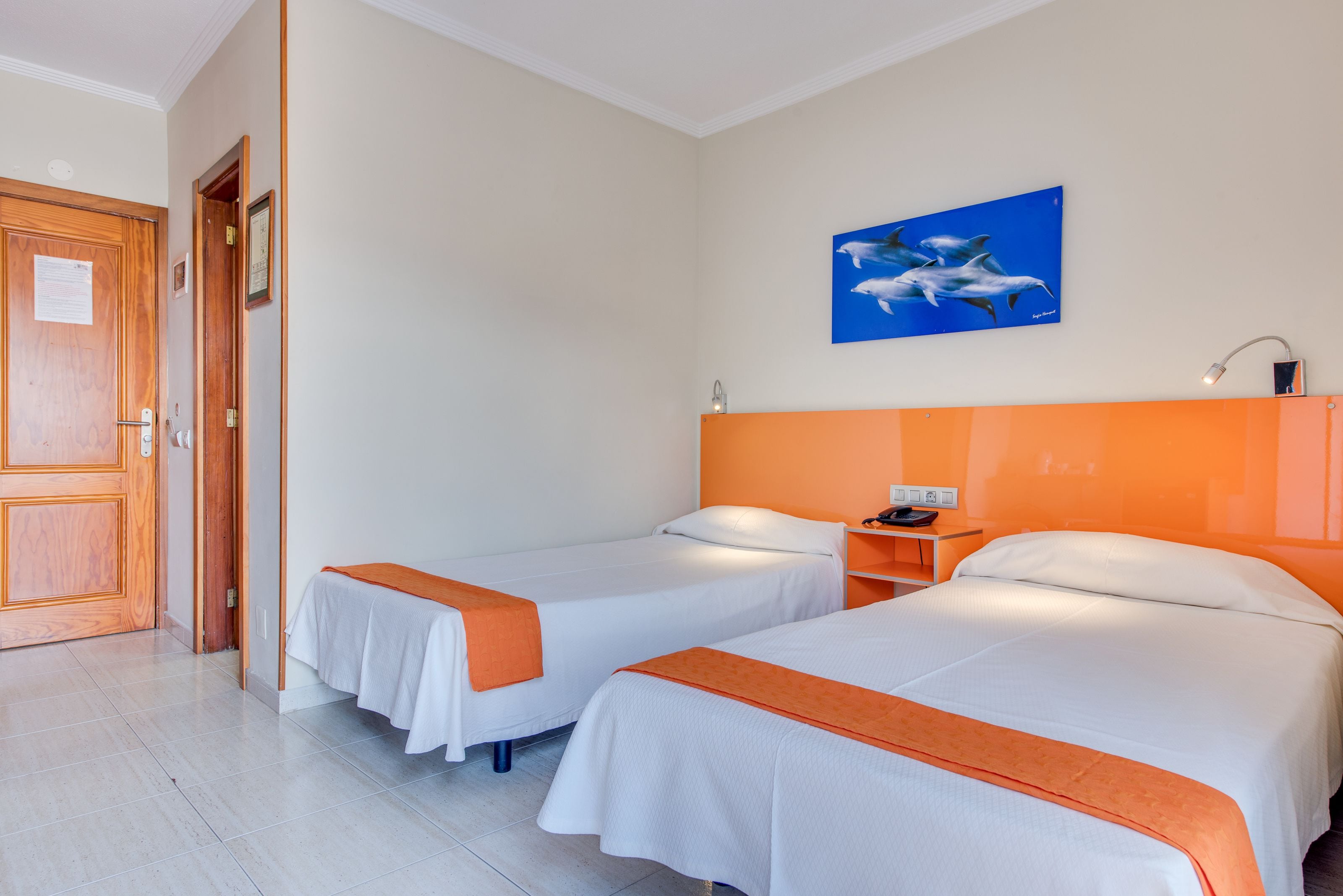 Quarto de hotel com duas camas, porta de madeira, pintura de golfinhos e mesa de cabeceira com abajur.