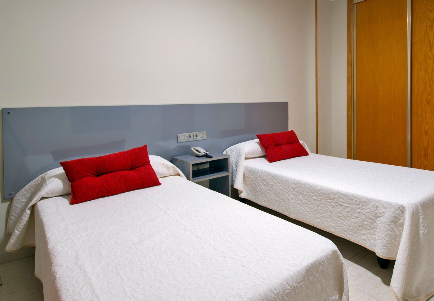 Habitación de hotel con dos camas individuales, cada una con almohada roja y ropa de cama blanca. Hay una mesita de noche con teléfono y toma de corriente.