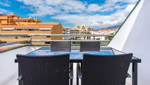 Un balcón con una mesa de vidrio y dos sillas de mimbre, con vistas a una hilera de edificios y un cielo despejado.