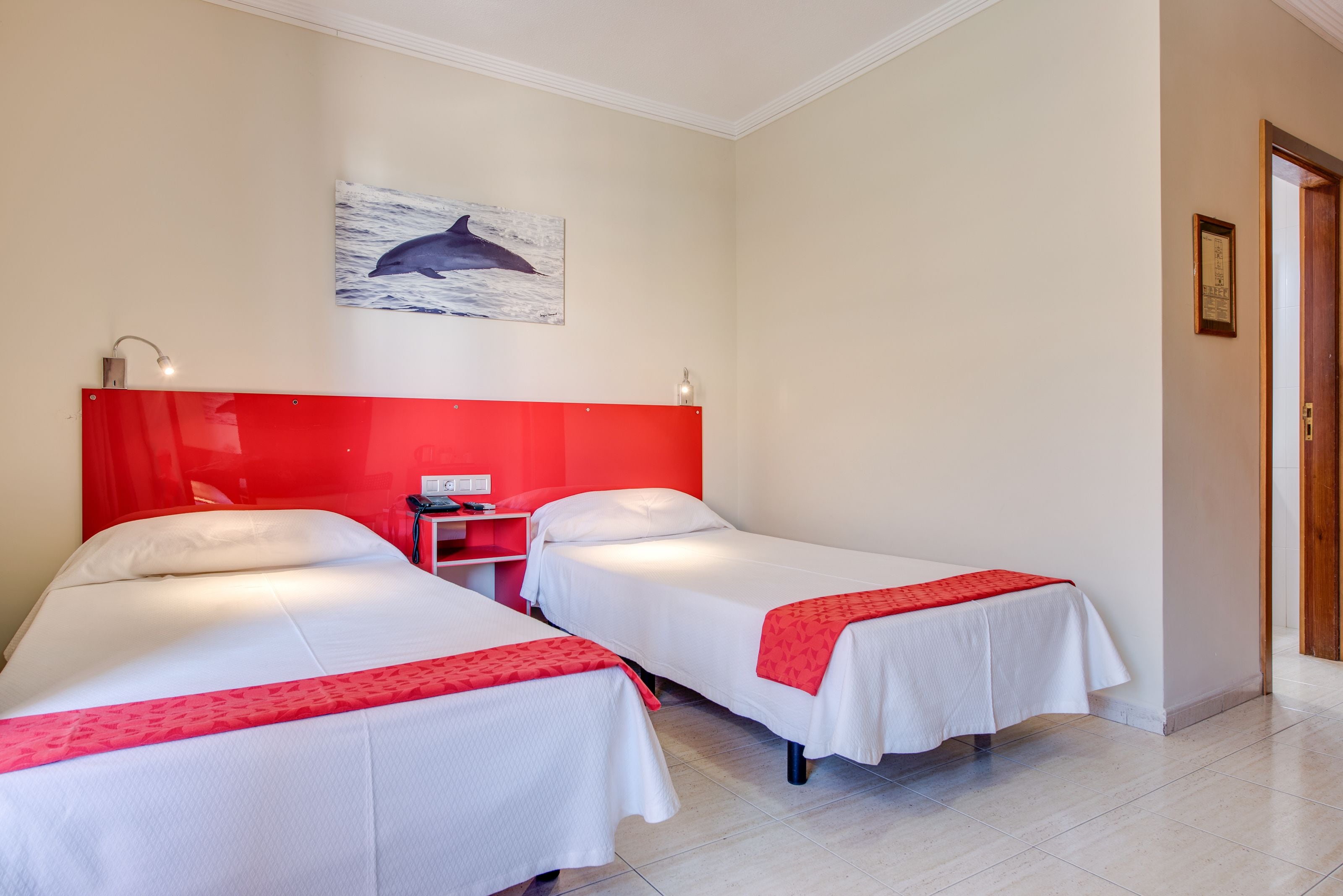 Habitación de hotel con dos camas, un cabecero rojo, un cuadro de delfín y un escritorio pequeño con una lámpara.