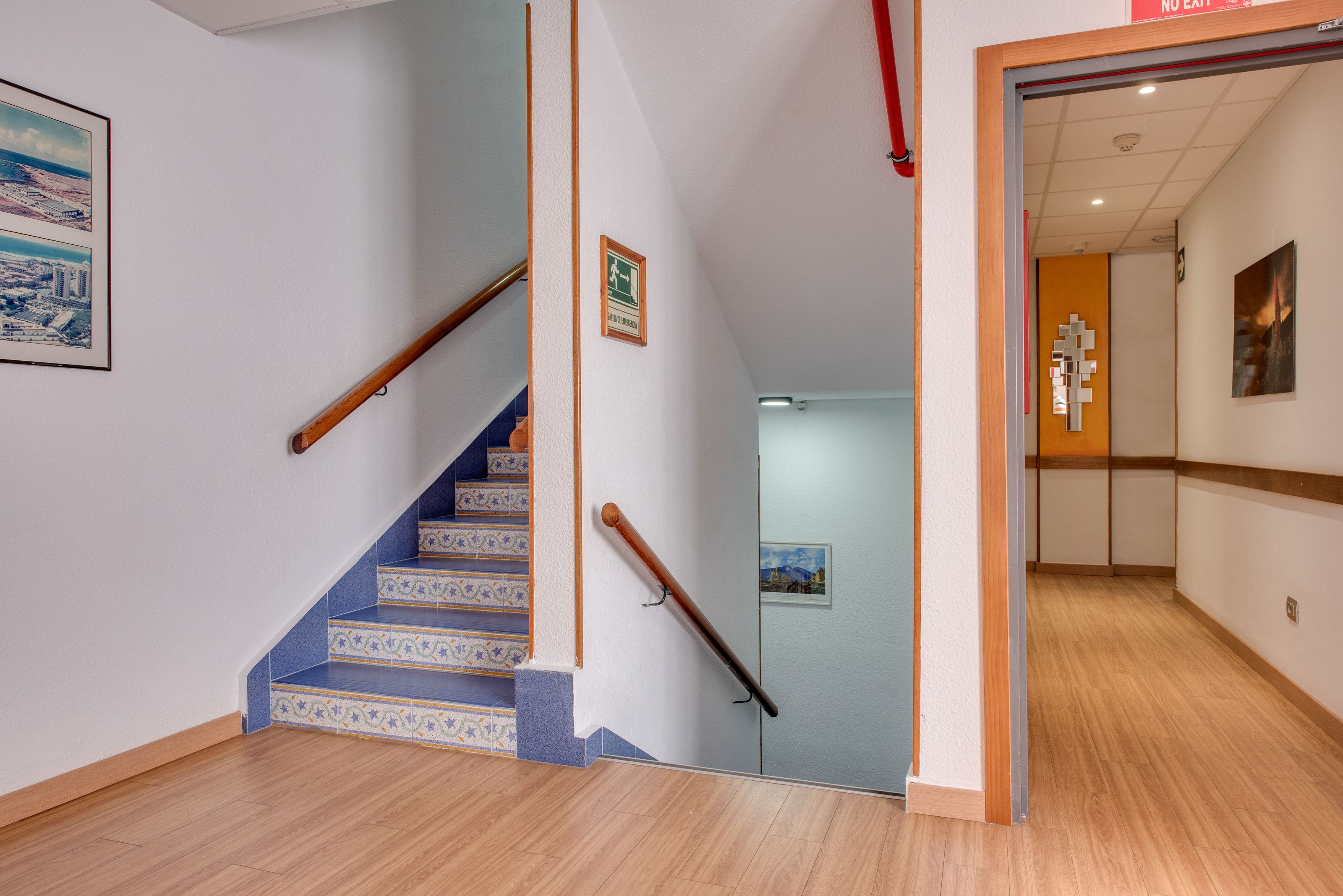 A staircase with blue patterned steps leading to a hallway with wooden flooring and a door with a red sign.