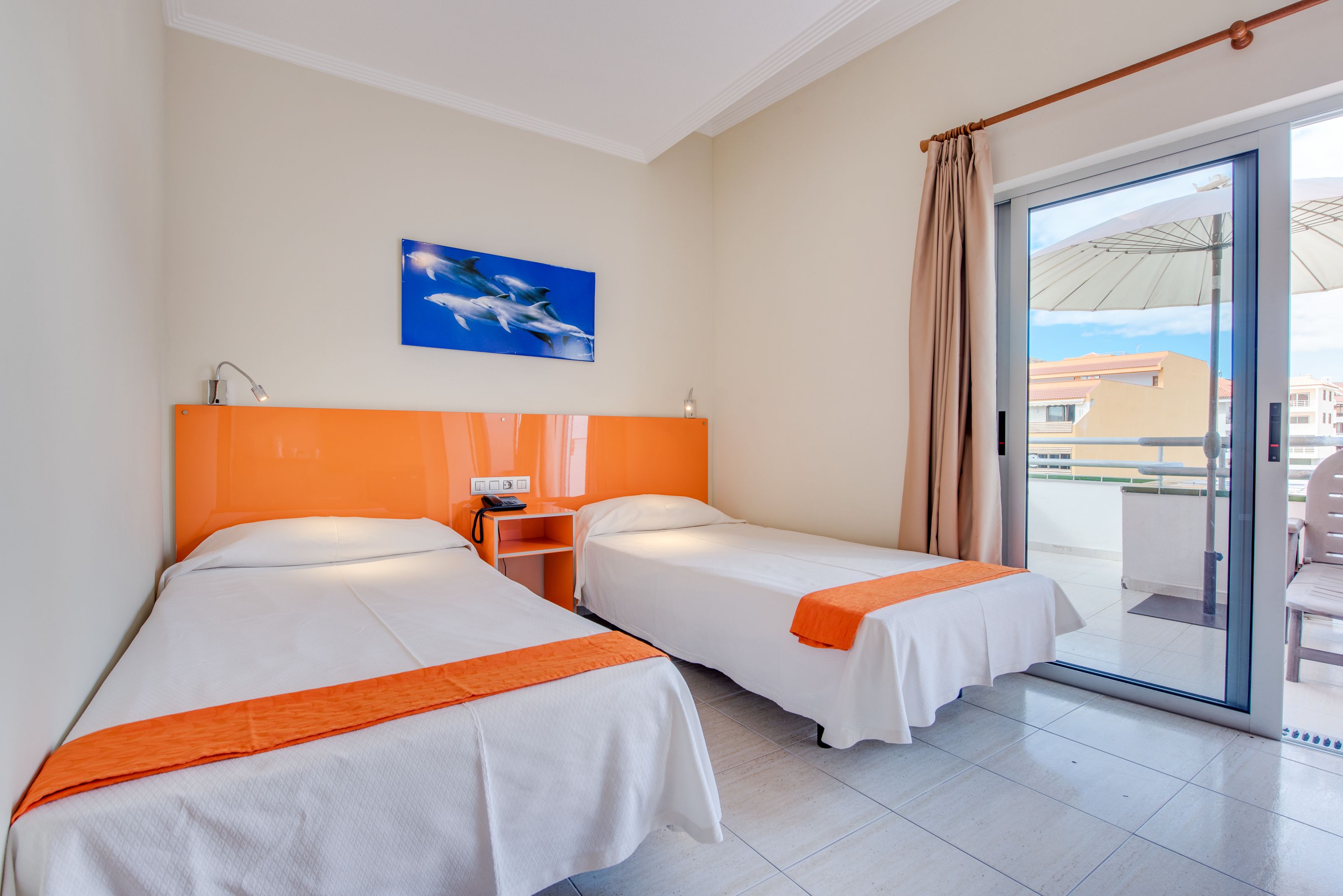 Habitación de hotel con dos camas, un cuadro de delfines, un balcón con vistas y una puerta corrediza de vidrio.