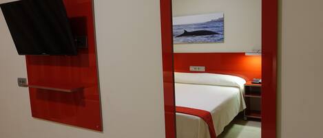A hotel room with a bed, a TV mounted on a red panel, a painting of a whale on the wall, and a red-framed mirror.