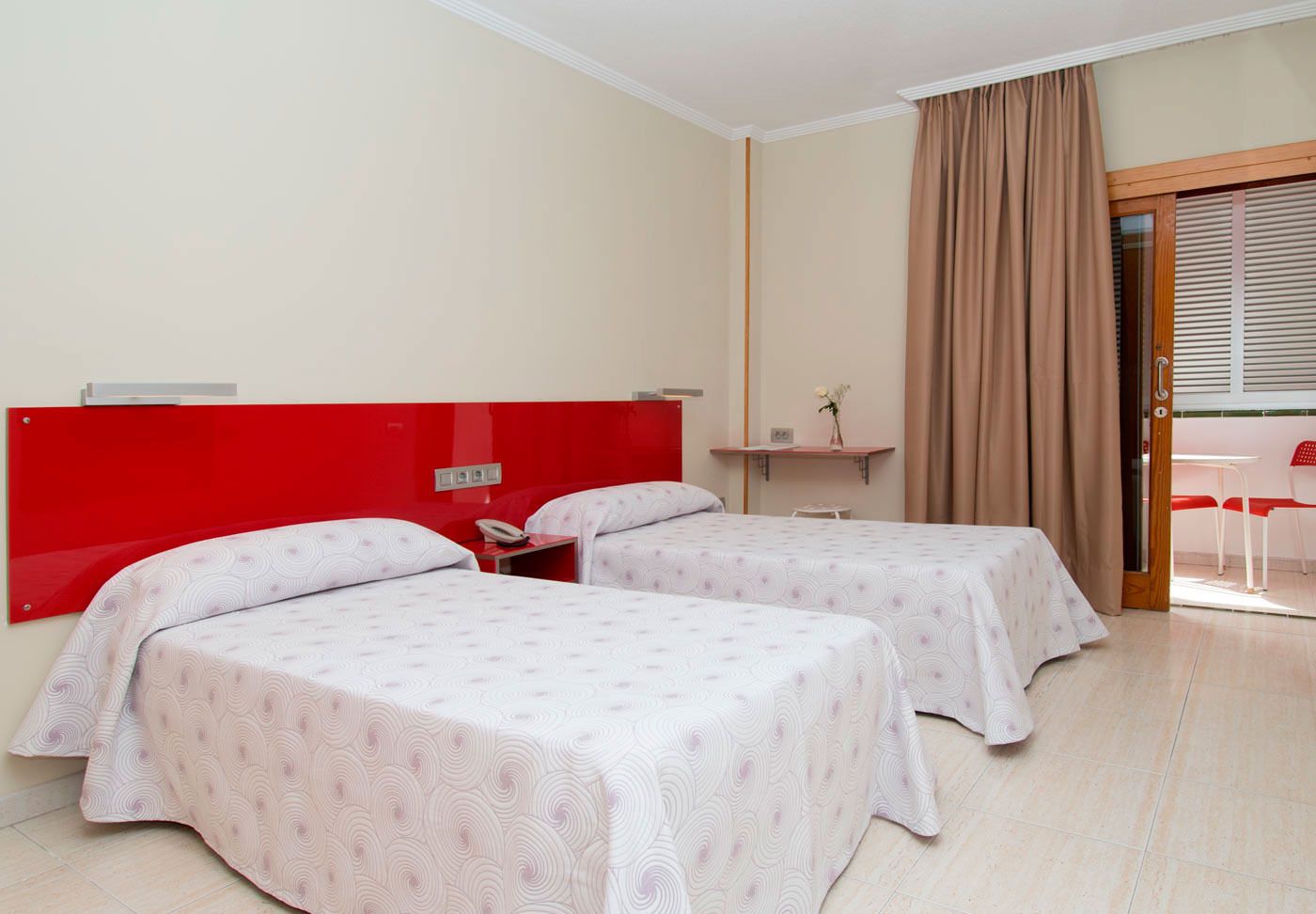 A hotel room with two beds, a red headboard, a small table with a phone, a window with blinds, and a door leading to a balcony.