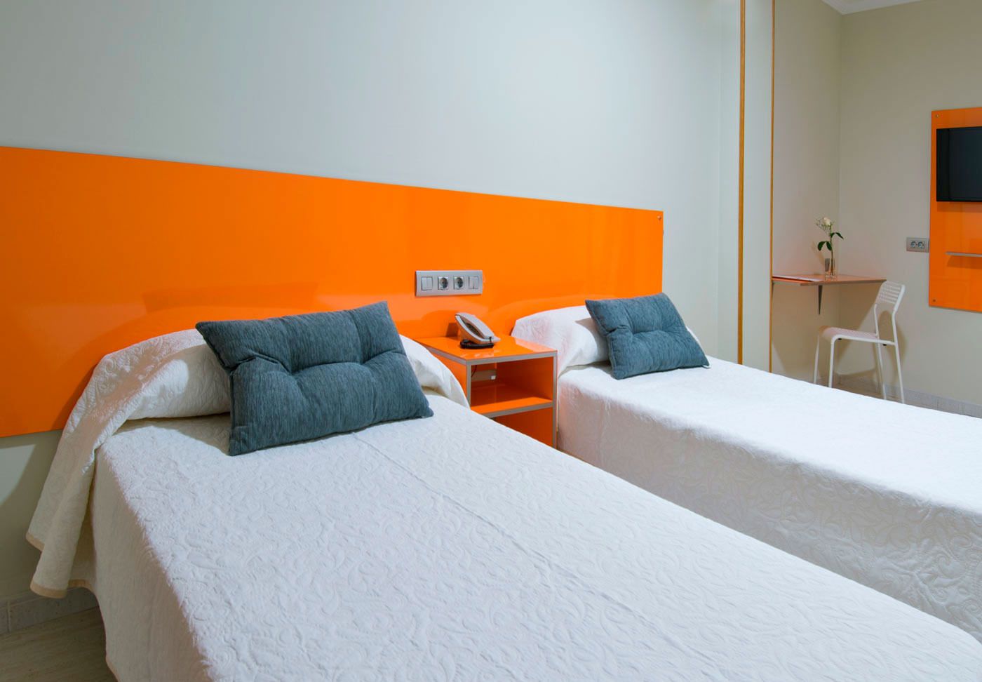 A hotel room with two beds, each with white bedding and blue pillows, a small table with a phone, and a chair.