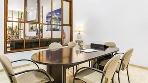 Un salón de conferencias con una mesa grande, cuatro sillas, una lámpara y vistas a una zona de estar a través de un panel de vidrio.