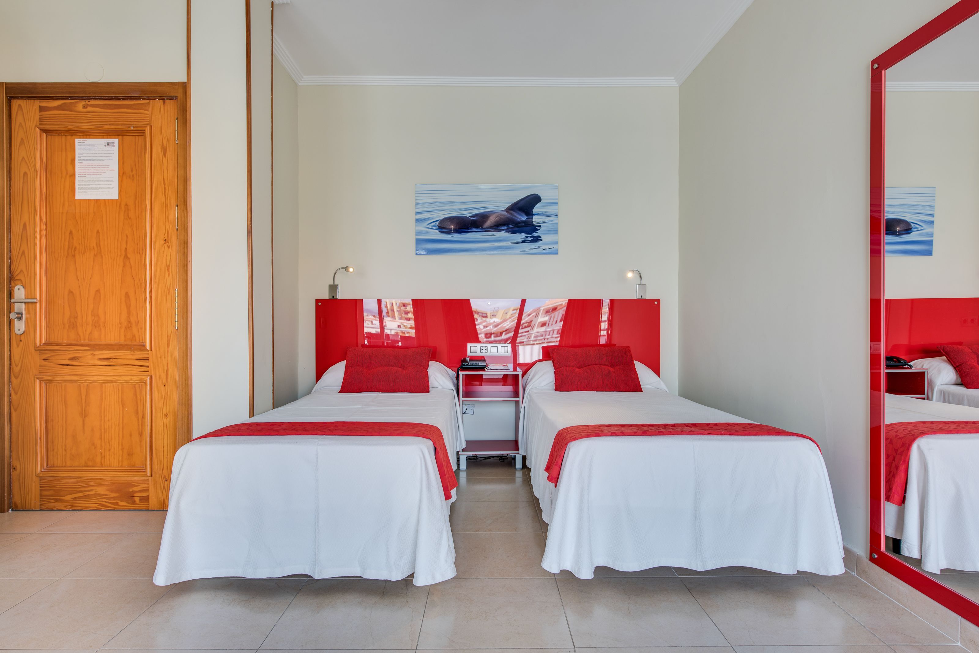 Habitación de hotel con dos camas, una puerta de madera, un cuadro de una ballena y un espejo con marco rojo.