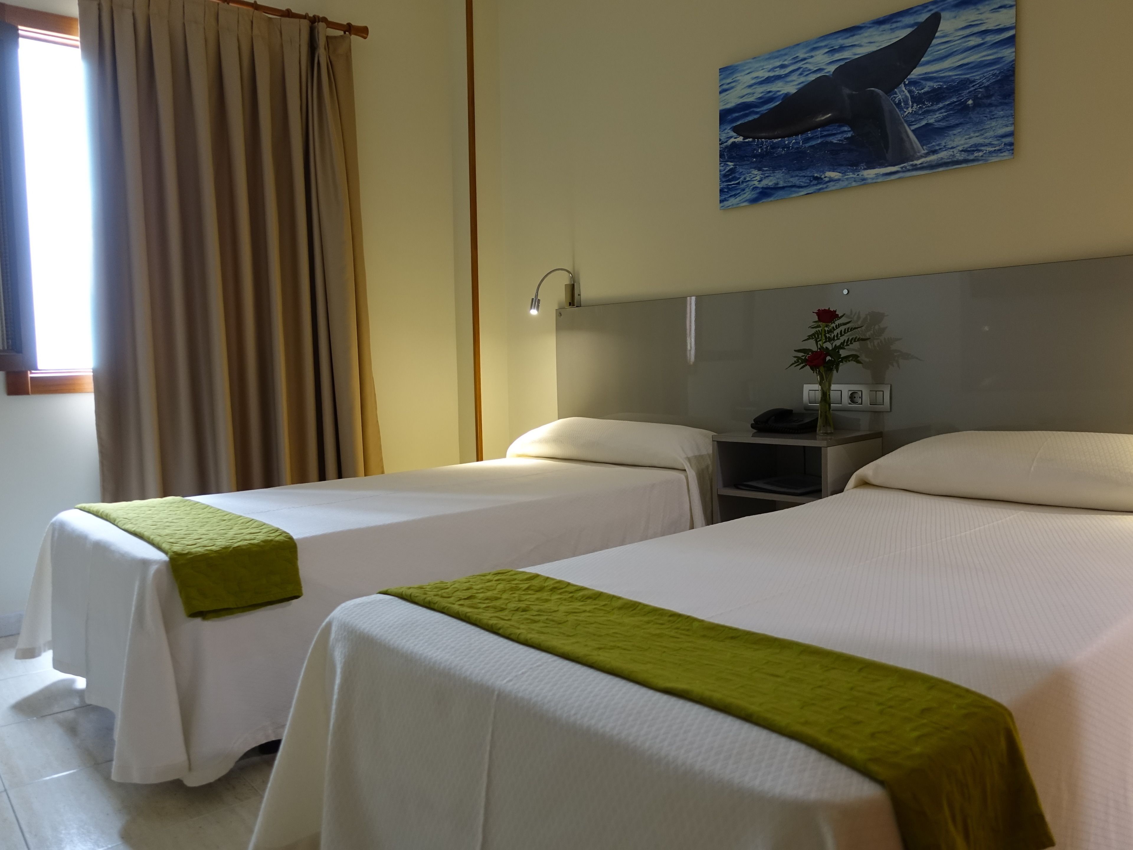 Habitación de hotel con dos camas, una mesita de noche con teléfono y un florero, un cuadro de ballena y una ventana con cortinas.