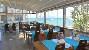 Een eetgedeelte met gedekte tafels, grote ramen met uitzicht op zee en een veraf gelegen eiland, en een hoge plafond met natuurlijk licht.