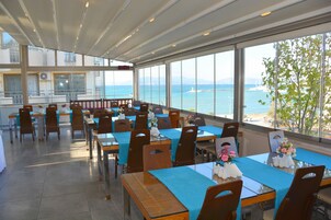 Un'area ristorazione con tavoli apparecchiati, ampie finestre con vista sul mare e un'isola sullo sfondo, e un soffitto alto con luce naturale.