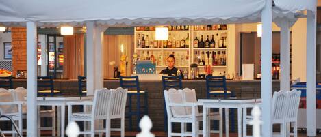 A bar area with white chairs and tables, a pool, and a bar stocked with bottles.