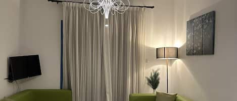 En moderne stue med to grønne sofaer, et hvidt sofabord, et fladskærms-tv, en gulvlampe og en lysekrone.