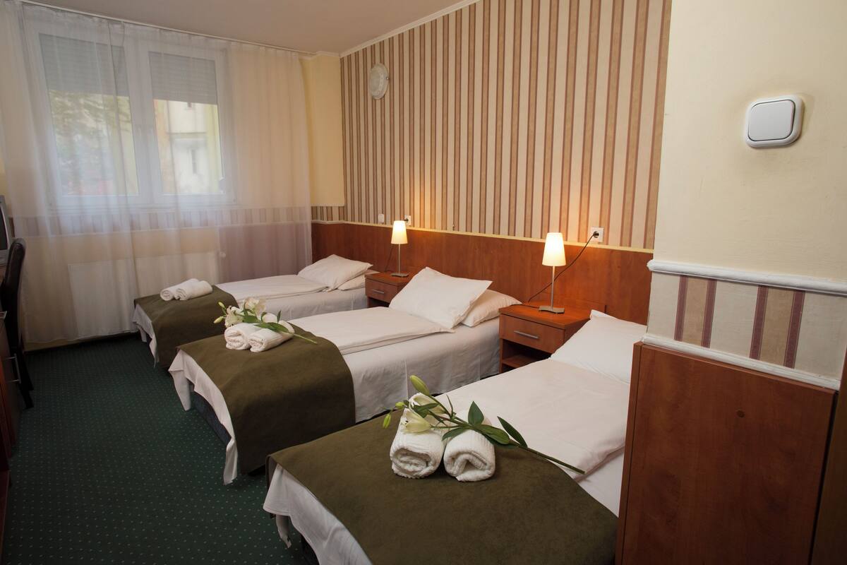 Hotellihuoneessa on kolme erillistä sänkyä, jokaisessa on tyyny ja pyyhe. Huoneessa on yöpöydät, joissa on lamput, seinällä on kello ja ikkunassa on läpikuultavat verhot.