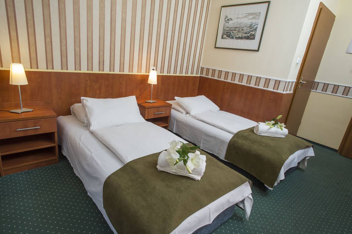 Hotellihuoneessa on kaksi sänkyä, joissa kummassakin on vihreä peitto ja valkoiset tyynyt, sekä pieni pöytä, jonka päällä on lamppu.