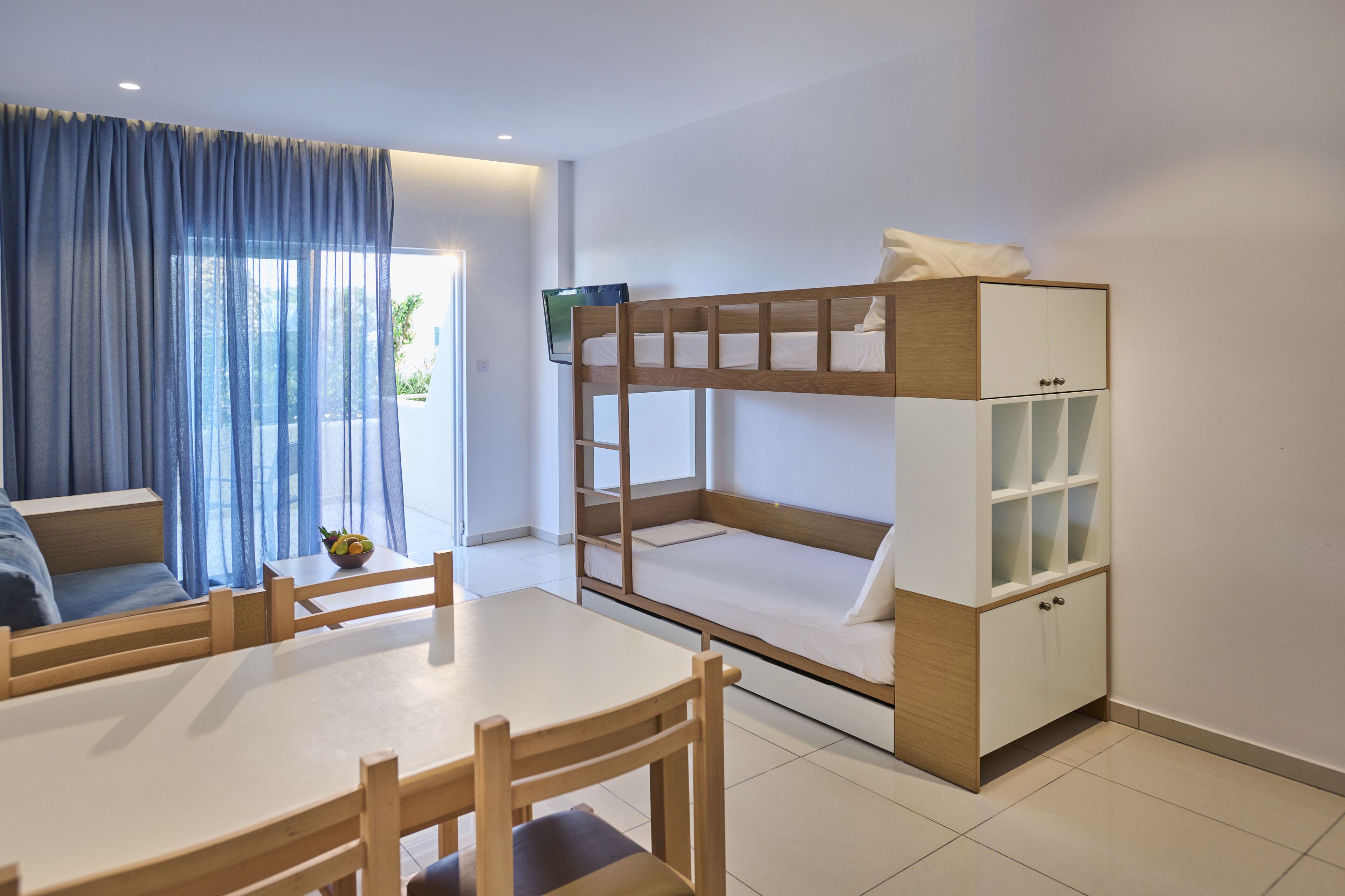 A compact room with a bunk bed, a small table with chairs, a sofa, and a cabinet with glass doors.