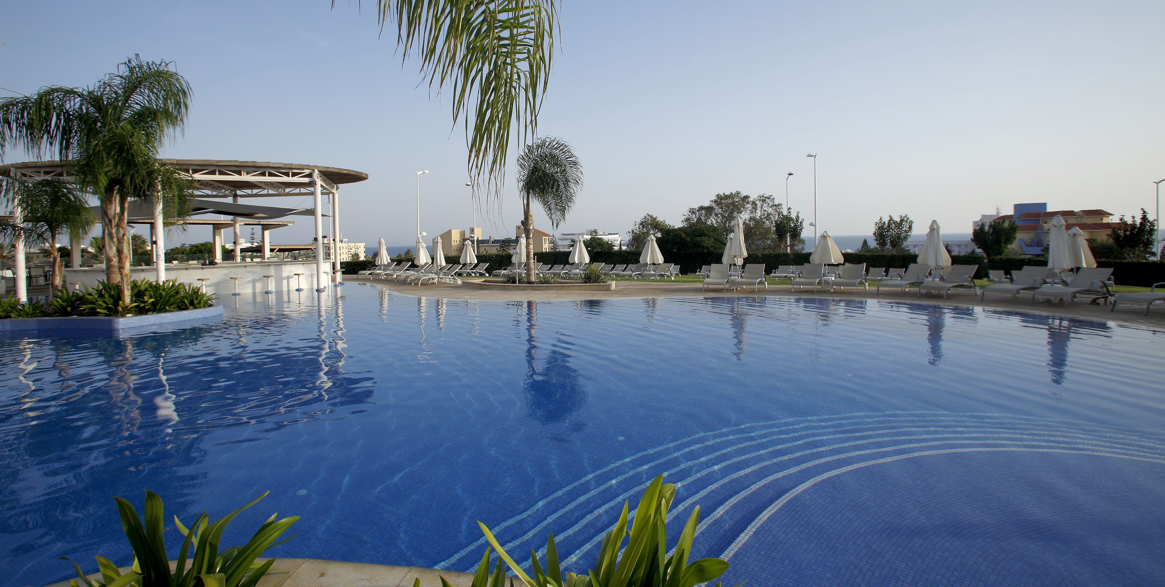 A swimming pool with lounge chairs and umbrellas, surrounded by palm trees and a covered seating area.