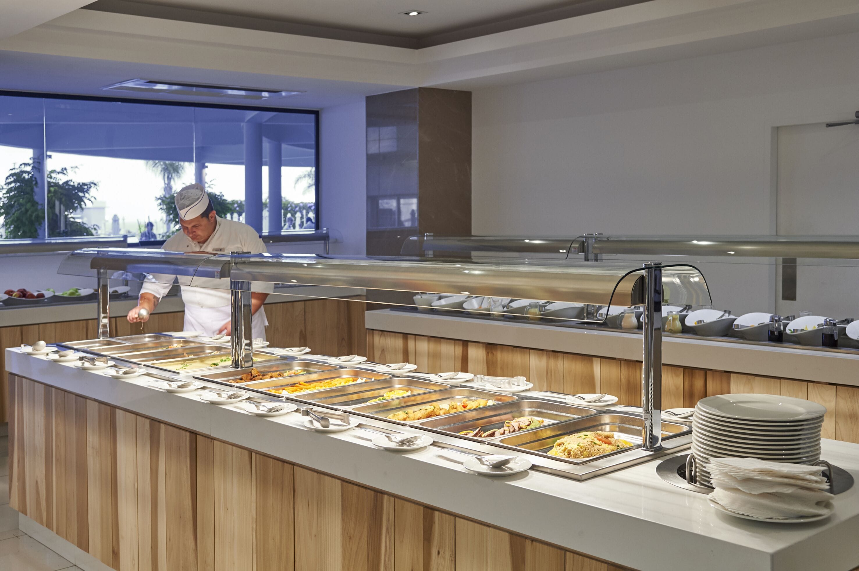 A buffet setup with multiple food stations, a counter with stacked plates, and a person serving food.