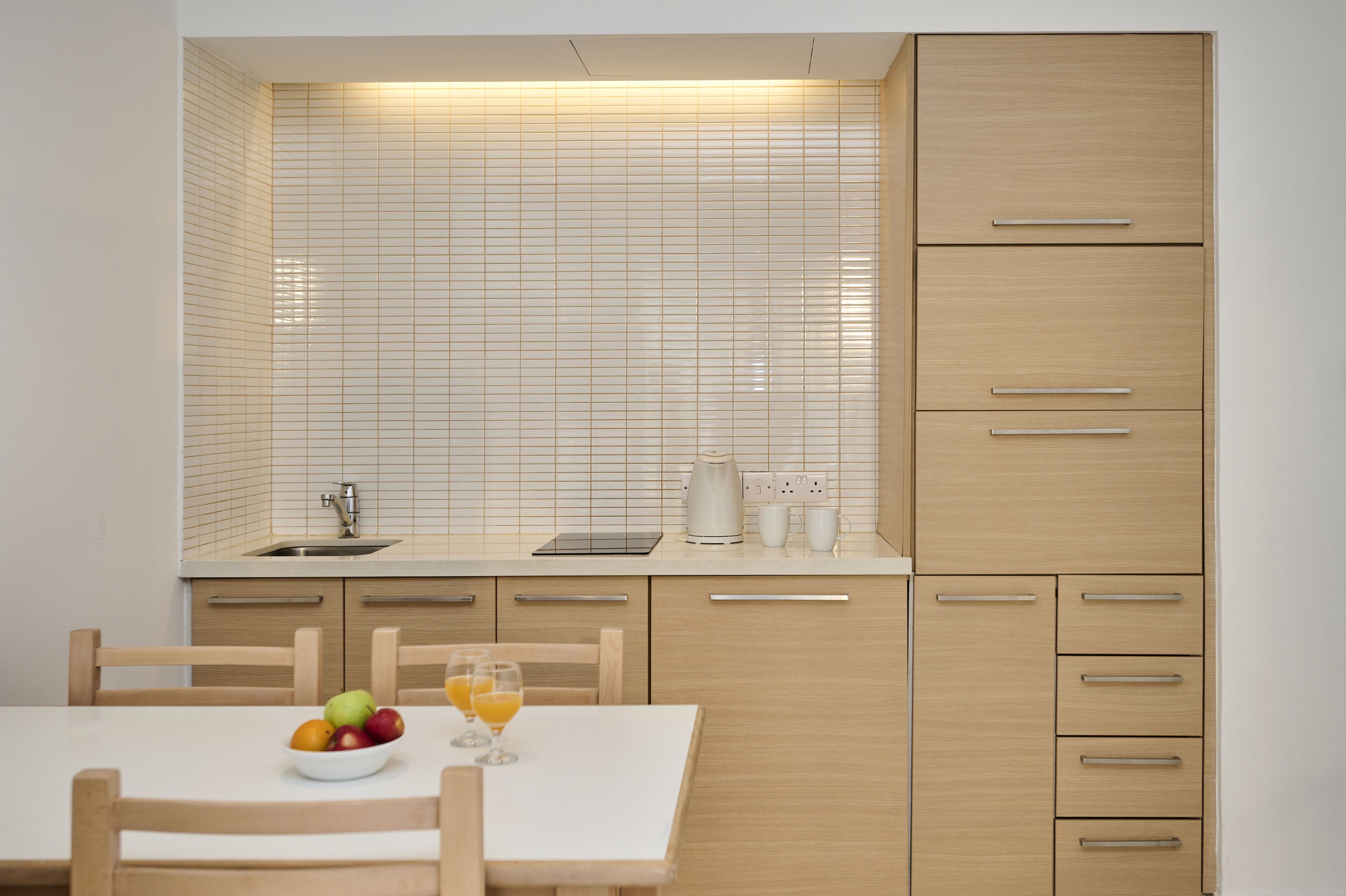 A modern kitchen with a white tiled backsplash, wooden cabinets, and a dining table with fruit and glasses.