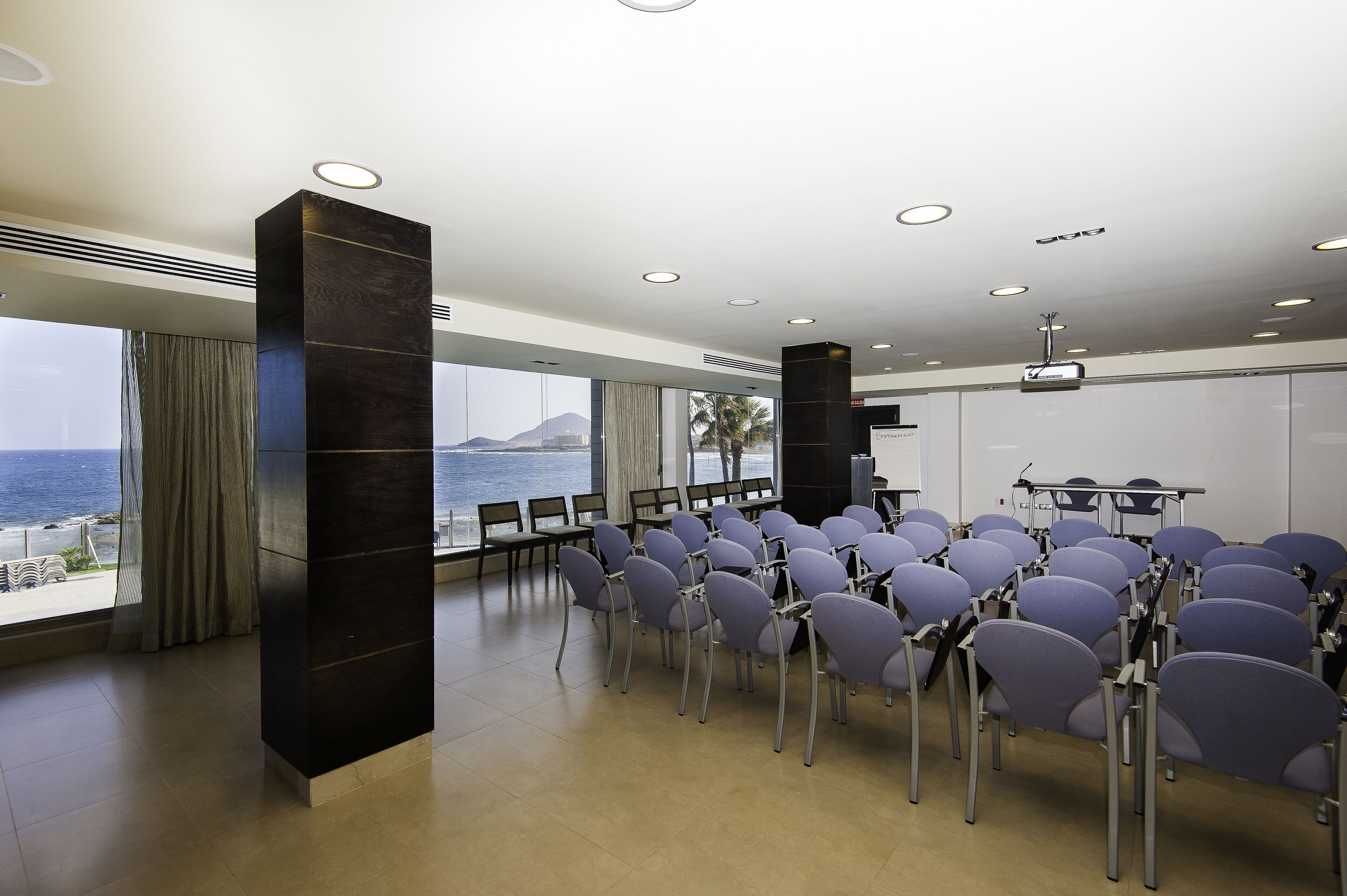 A conference room with a view of the sea, rows of chairs, a projector, and a large pillar.