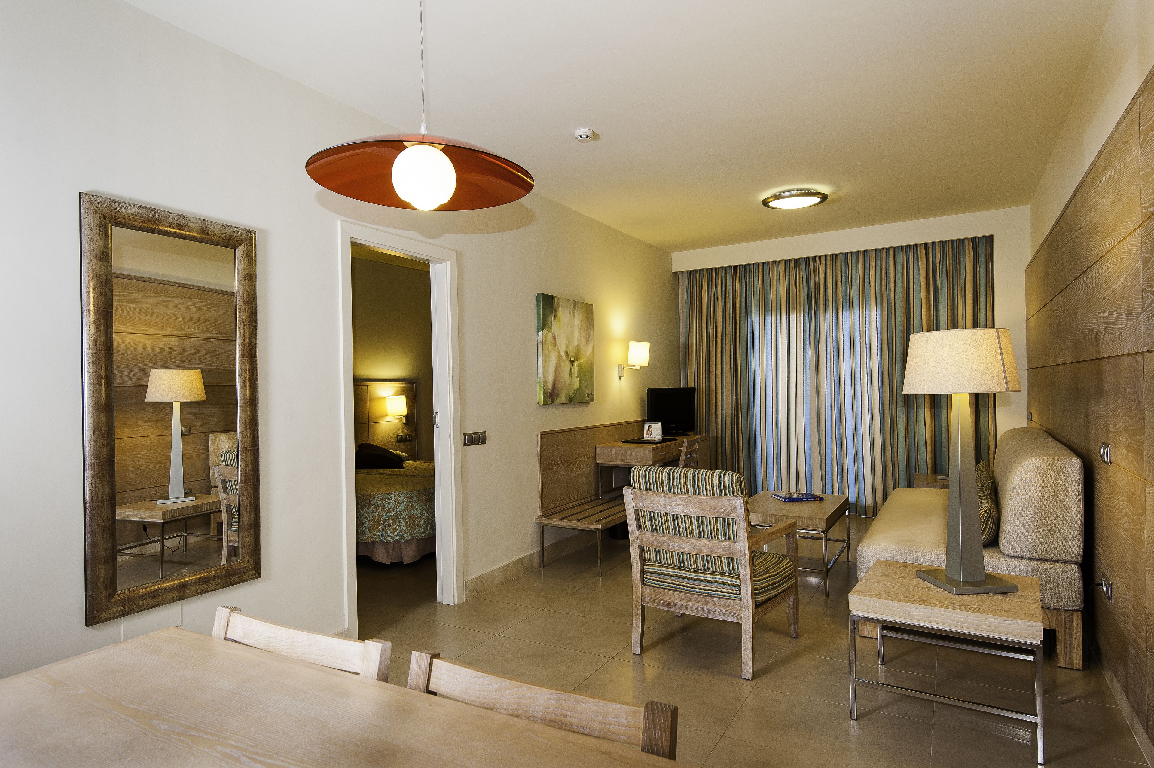 Una habitación de hotel moderna con comedor, sofá, televisión y un amplio ventanal con cortinas.